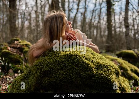 Snowdrops galanthus blond. Une fille en robe blanche s'est couché sur une pierre dans la mousse dans un pré avec des gouttes de neige dans une forêt de printemps Banque D'Images