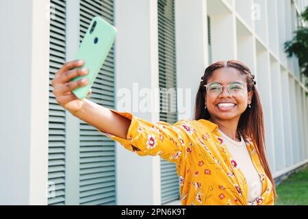 Une étudiante afro-américaine souriant prenant un portrait de selfie avec un téléphone portable, montrant un sourire crasseux tout en se tenant debout à l'extérieur du campus de l'université. Adolescent positif prenant photo avec téléphone mobile . Photo de haute qualité Banque D'Images