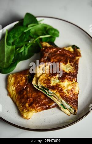 Omelette pliée farcie aux épinards et à la feta sur l'assiette blanche avec des feuilles d'épinards au dos. Plat de petit déjeuner sain et diététique. Cuisine minimaliste Banque D'Images