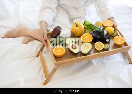 Vue rapprochée de la femme dans un peignoir tenant le plateau de lit avec des fruits frais et des produits de soin du visage tout en étant assis sur une couverture blanche. Ingrédients naturels pour le soin du visage disponibles à la maison. Banque D'Images