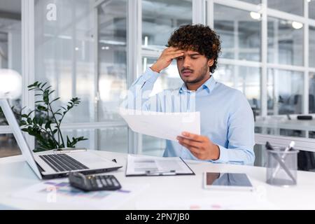 Financier contrarié regardant les ventes et le tableau des revenus de l'entreprise, homme d'affaires au sein du bureau déçu par les résultats et les réalisations des ventes, homme hispanique assis à table avec des documents et un ordinateur portable. Banque D'Images