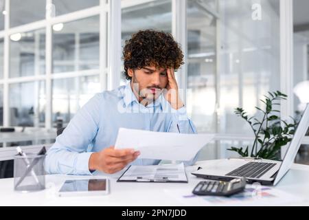 Financier contrarié regardant les ventes et le tableau des revenus de l'entreprise, homme d'affaires au sein du bureau déçu par les résultats et les réalisations des ventes, homme hispanique assis à table avec des documents et un ordinateur portable. Banque D'Images