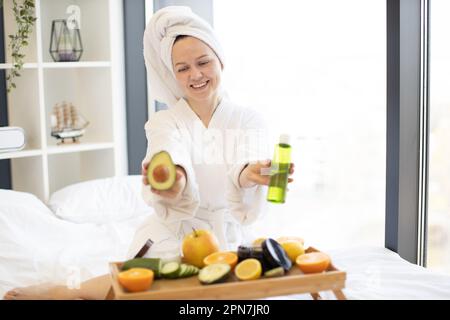 Femme souriante dans une tenue après-douche présentant l'avocat vert aux fruits et à l'huile cosmétique tout en étant assise derrière le plateau de lit à la maison. Une dame joyeuse recommande des ingrédients naturels dans la thérapie du visage. Banque D'Images