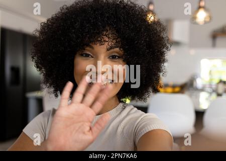 Portrait d'une heureuse femme biraciale avec aro assis à la maison, faisant des appels vidéo, agitant et souriant Banque D'Images