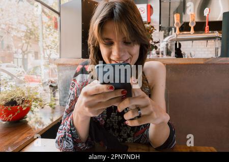 Vue de face de la jeune femme transgenre hispanique latino-argentine avec de longs cheveux, souriant heureux en utilisant le téléphone assis dans un restaurant, en attendant Banque D'Images