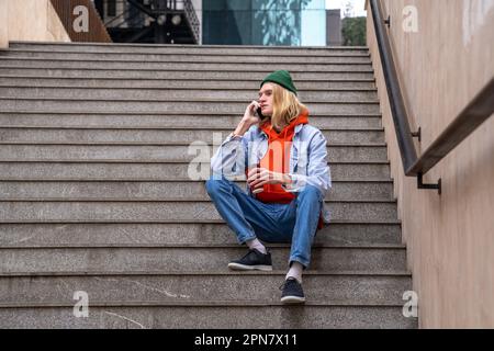 Un homme élégant avec une tasse de café à la main s'assoit sur des marches parle sur un smartphone dans la rue de la ville. Banque D'Images