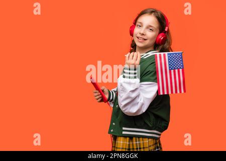 Une jeune fille d'école en vêtements décontractés et écouteurs tient un petit drapeau américain, ou le drapeau américain et smartphone. Cours de langues en ligne. Apprendre Ameri Banque D'Images