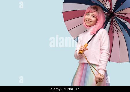 Portrait adolescent avec un autre look sur fond bleu studio. Jeune fille avec perruque de cheveux roses et parapluie multicolore, porte le clothe de couleur pastel Banque D'Images