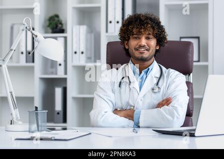 Portrait d'un docteur hindou réussi dans le bureau d'une clinique moderne, assis sur le lieu de travail à la table souriant et regardant la caméra avec ses bras croisés, un homme dans un manteau blanc. Banque D'Images
