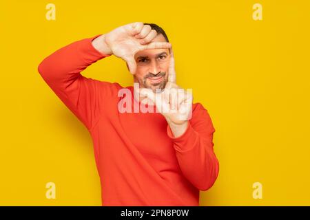 Homme hispanique barbu dans son 40s portant un chandail rouge faisant un cadre avec ses doigts, isolé sur fond jaune. Concept de créativité. Banque D'Images