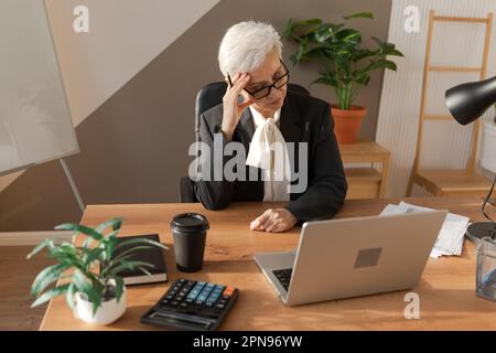 Femme âgée touchant des temples éprouvant du stress au bureau. Femme mature fatiguée du travail de sentir mal de tête de frottement des temples malades front. Utilisation prolongée d'un ordinateur portable syndrome de vision d'ordinateur. Syndrome de bureau Banque D'Images