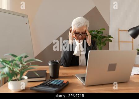 Femme âgée touchant des temples éprouvant du stress au bureau. Femme mature fatiguée du travail de sentir mal de tête de frottement des temples malades front. Utilisation prolongée d'un ordinateur portable syndrome de vision d'ordinateur. Syndrome de bureau Banque D'Images