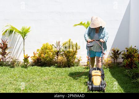 Femme portant un short bleu, une chemise à manches longues et un chapeau, tond l'herbe dans le jardin de sa maison avec une tondeuse à gazon. Banque D'Images