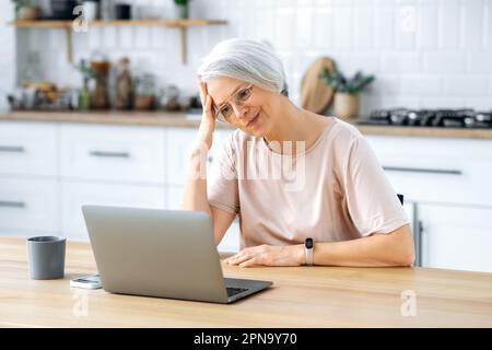 Insatisfait de la femme d'âge moyen de race blanche avec des cheveux gris, portant des lunettes, assis à une table avec un ordinateur portable dans la cuisine à la maison, travaille à distance, regarde l'écran dans la confusion, lit les nouvelles Banque D'Images