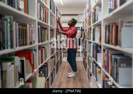 Homme étudiant afro-américain réfléchi dans des verres choisir le livre dans la bibliothèque d'université ou la librairie Banque D'Images