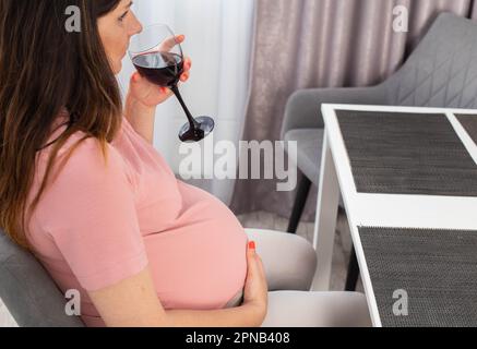 Une femme enceinte s'assoit dans la cuisine à la table et boit de l'alcool. Mauvaise habitude pendant la grossesse. Banque D'Images