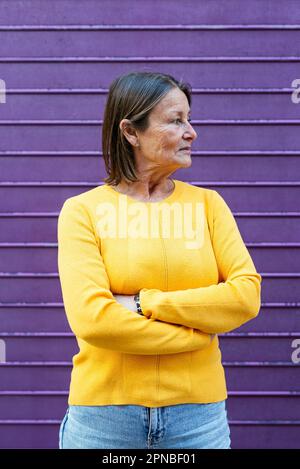 Femme d'âge moyen dans des vêtements jaune vif regardant loin tout en se tenant contre le mur violet Banque D'Images