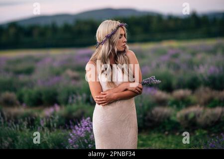 Jeune femme en robe blanche debout dans un champ de lavande en fleur avec les yeux fermés appréciant la nature pendant le coucher du soleil Banque D'Images