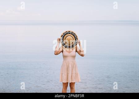 Une femme voyageur anonyme couvrant la face avec un chapeau de paille tout en se reposant sur la côte d'un océan calme et sans fin, par temps sombre et nuageux Banque D'Images