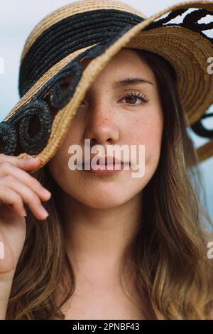 Portrait d'une femme modèle couvrant la face avec chapeau de paille et regardant l'appareil photo tout en se reposant sur les vacances d'été sur fond bleu Banque D'Images