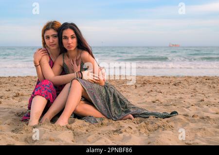 Ensemble complet de jeunes filles attentionnés dans de longues robes assis sur une plage de sable près de la mer et de s'embrasser tout en regardant l'appareil photo Banque D'Images