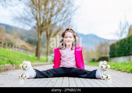 Fille positive enfant assis avec des jambes étirées sur une passerelle en bois et se détendre tout en regardant la caméra pendant le patinage avec patins à roulettes dans le parc i Banque D'Images