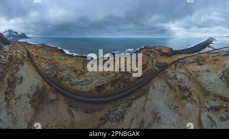 Vue par drone des vagues lavant la côte rocheuse avec la route asphaltée traversant un terrain montagneux pendant la journée en Islande Banque D'Images