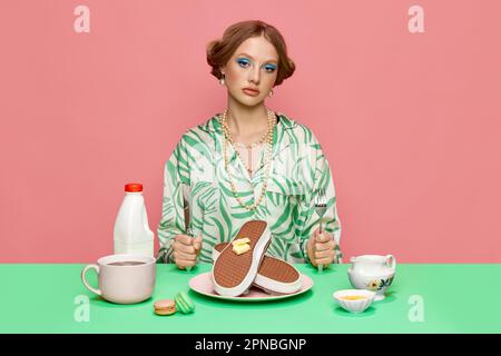 Portrait d'une jeune fille jolie et élégante assise à table et mangeant des sneakers avec gaufres au beurre pour le petit déjeuner sur fond rose studio Banque D'Images