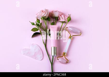 Rouleau de jade rose et le gua sha pour le massage du visage avec de la crème ou du gel pour le visage et les roses de fleur. Outils de massage pour le visage. Véhicule anti-âge, levage et virage Banque D'Images