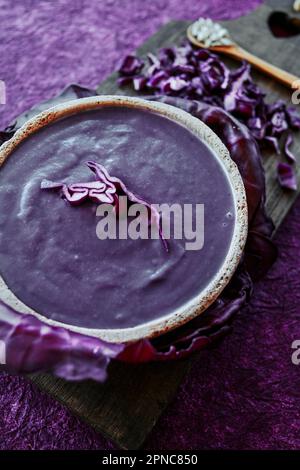 gros plan d'une soupe de chou rouge dans un bol en céramique, placé sur une planche à découper en bois foncé, sur une surface texturée mauve Banque D'Images