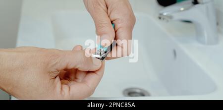 un homme coupe ses ongles avec un coupe-ongles, en appuyant sa main sur le lavabo blanc de sa salle de bains, dans un format panoramique à utiliser comme bannière web Banque D'Images