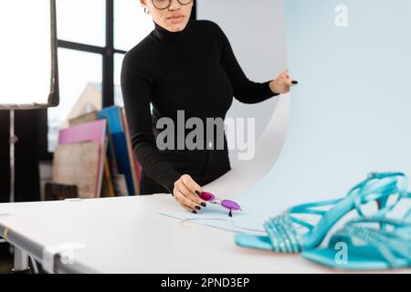 vue rognée du fabricant de contenu afro-américain tenant des lunettes de soleil à la mode près des sandales floues sur la table de tournage dans le studio photo, image de stock Banque D'Images