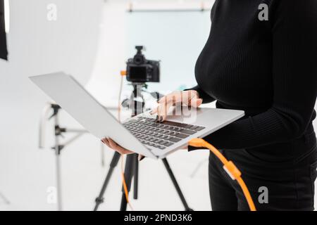 vue rognée du gestionnaire de contenu afro-américain à l'aide d'un ordinateur portable près d'un appareil photo numérique flou sur trépied en studio photo, image de stock Banque D'Images