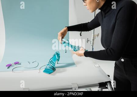 vue partielle du gestionnaire de contenu afro-américain tenant la chaussure à la mode près de lunettes de soleil et des accessoires de bijoux sur la table de tournage dans le studio photo, stock i Banque D'Images