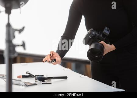 vue partielle d'un photographe afro-américain avec un appareil photo numérique tenant un mascara près des ombres à paupières et des pinceaux cosmétiques sur une table de tournage en studio, st Banque D'Images