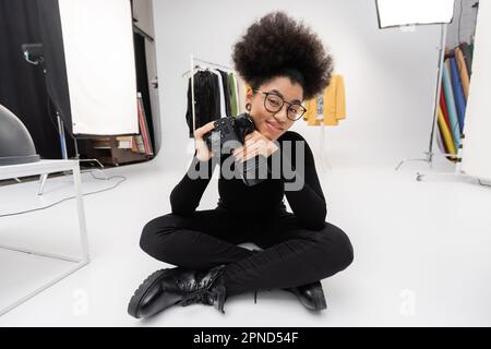 femme afro-américaine souriante vêtue de noir assis avec un appareil photo numérique professionnel dans un studio photo moderne, stock image Banque D'Images