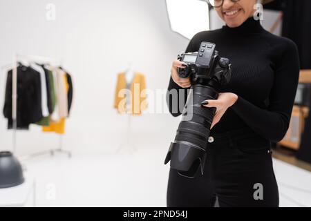 vue partielle d'un joyeux photographe afro-américain en col de cygne noir tenant un appareil photo numérique professionnel en studio photo flou, image de stock Banque D'Images