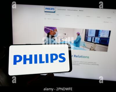 Personne tenant un téléphone portable avec le logo de la société néerlandaise Koninklijke Philips N.V. à l'écran en face de la page Web d'affaires. Mise au point sur l'affichage du téléphone. Banque D'Images