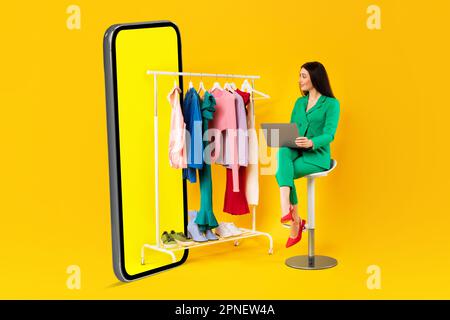 Femme emballée designer de mode avec ordinateur portable regardant des vêtements sur rail près de l'énorme téléphone portable sur fond jaune studio Banque D'Images