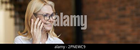 Femme d'âge moyen occupée avec des cheveux blonds et des lunettes dans une chemise blanche buvant du café ou du thé tout en parlant au téléphone, debout à la maison Banque D'Images