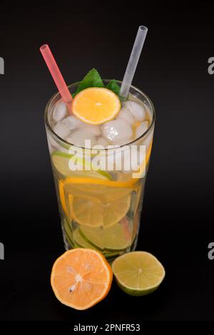 Limonade rafraîchissante dans un grand verre à facettes avec de la glace et des morceaux d'agrumes sur fond noir, à côté de la moitié d'un citron mûr et de lime. Disposition verticale Banque D'Images