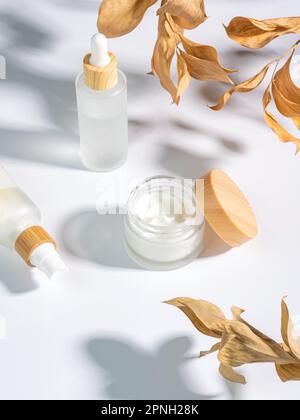 gros plan de la crème de collagène dans un pot en verre, du sérum dans un flacon en verre blanc mat et des feuilles de ruskus sèches sur fond blanc. Kit pour les soins de la peau et du corps Banque D'Images