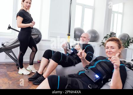 Un couple d'âge heureux en combinaisons ems s'assoit sur des chaises de massage après un entraînement de stimulation musculaire électrique avec un entraîneur personnel dans la salle de gym ou le club de fitness. Sourire Banque D'Images