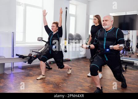 Un couple de personnes âgées dans des combinaisons ems faisant de l'exercice de stimulation musculaire électrique dans une salle de gym moderne avec un entraîneur personnel. Femme blonde plentue faisant des fentes et des squats Banque D'Images
