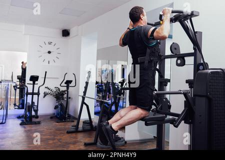 Un jeune homme en costume ems se met en mouvement sur un simulateur sportif. Des athlètes masculins tendus s'entraîne sur un appareil gravitron dans une salle de sport ou un club de fitness. Muscle électrique stimulateur Banque D'Images