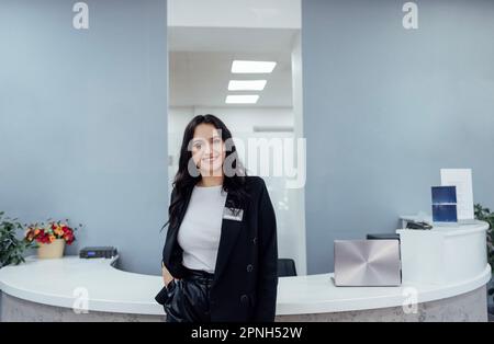 Une fille souriante de brune accueille à la réception dans un bureau moderne. La jeune femme en costume noir et en chemise blanche est debout près du comptoir semi-circulaire. PC portable, p Banque D'Images