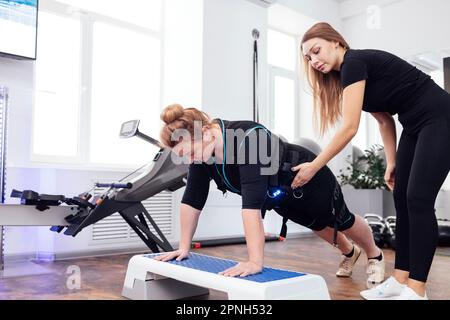 La femme d'âge moyen de Plump dans la combinaison ems est engagée sur la plate-forme de marche avec entraîneur personnel de forme physique. Femme blonde faisant des push-up du stepper. Électrique Banque D'Images