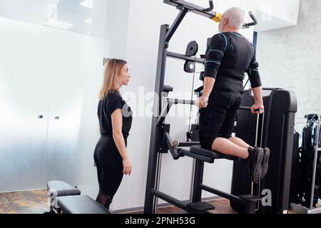 L'homme âgé aux cheveux gris en costume ems est engagé sur les barres inégales du simulateur gravitron avec des entraîneurs personnels en salle de gym. Alimentation électrique muscle sti Banque D'Images