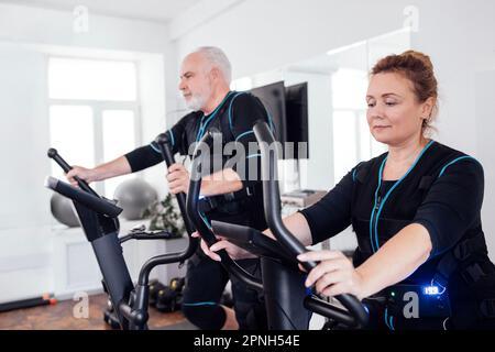 Deux personnes âgées en costumes ems sont engagées dans des équipements de sport. Le vieil homme fait de l'exercice sur un entraîneur elliptique. Femme âgée graisse exercice de pédalage b Banque D'Images