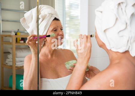 Mature belle femme appliquant le masque facial vert maison DIY sur la peau. Regarder dans le miroir dans la salle de bain, enveloppé dans une serviette. Routine de soin de la peau Banque D'Images